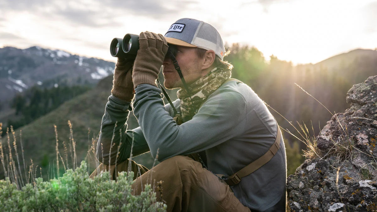 Kneeling person using dark green binoculars with a chest harness, overlooking mountains.