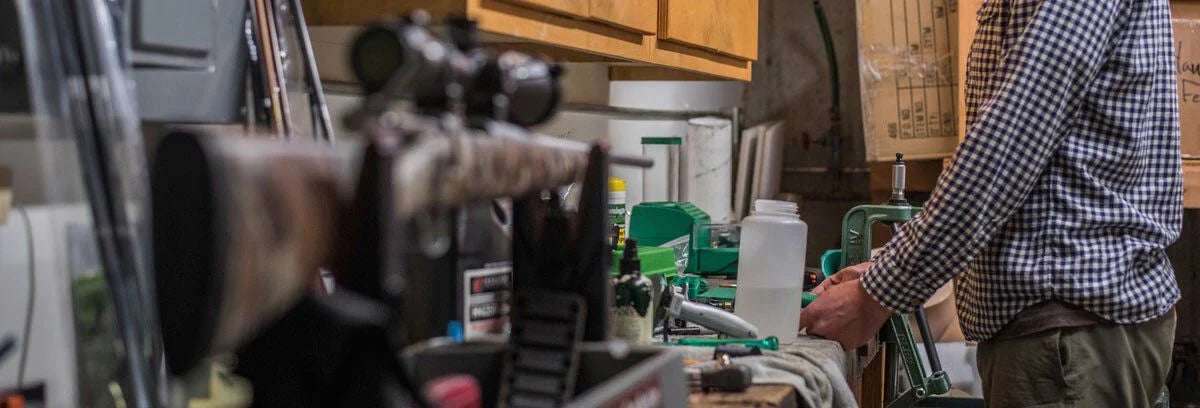 Person operates green reloading press in a workshop with a scoped camo rifle.