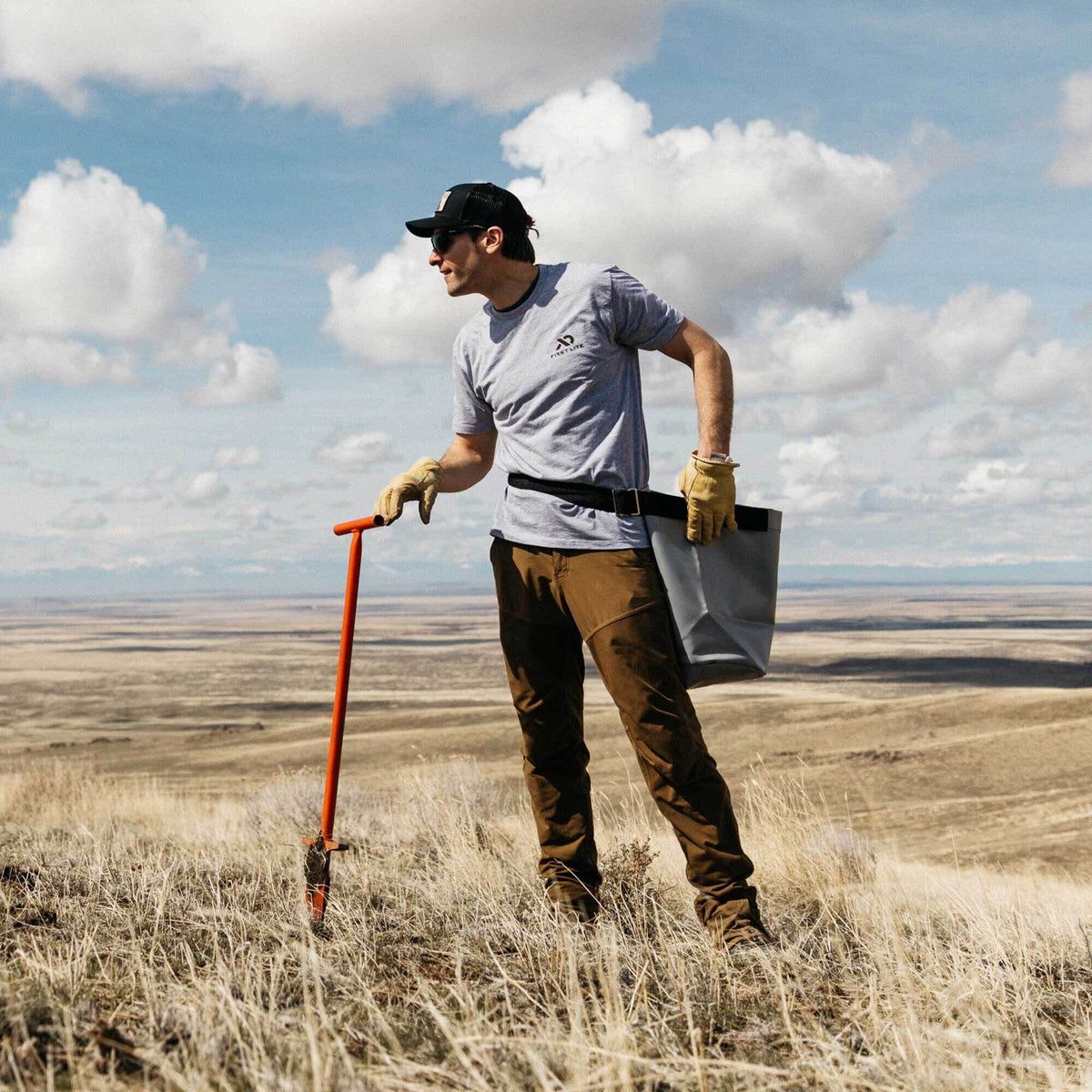 Man holding an orange L-shaped dibble bar and wearing a grey harvesting bag on a black belt around his waist.