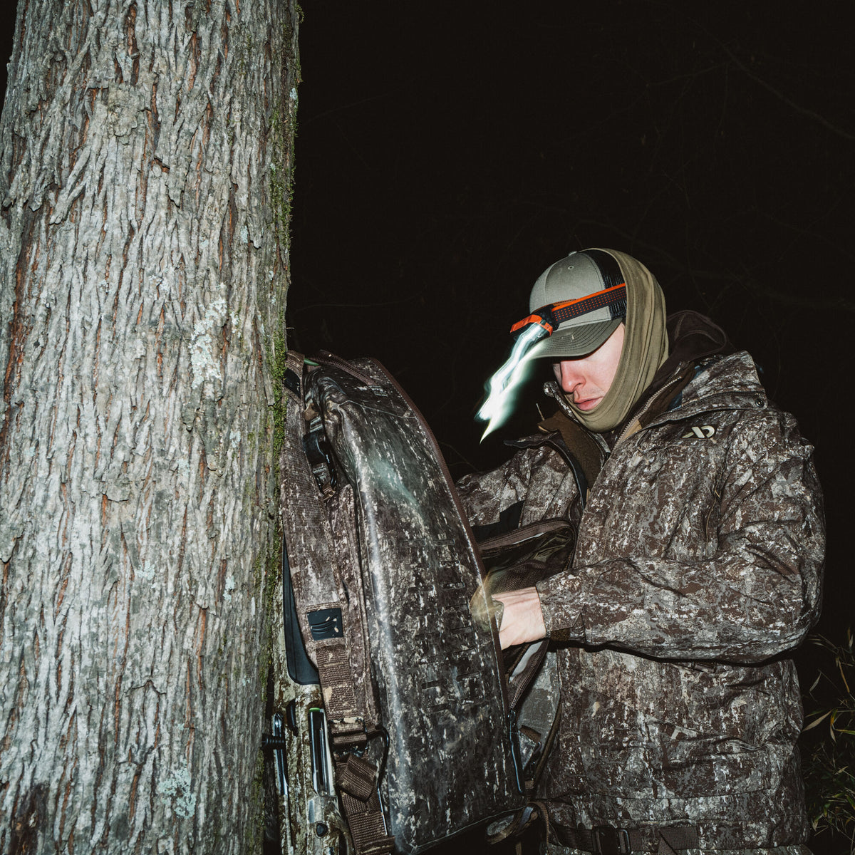 A brown and grey camouflage hunting gear bag with a rigid back panel is mounted on a tree, being adjusted by a person wearing a headlamp at night.