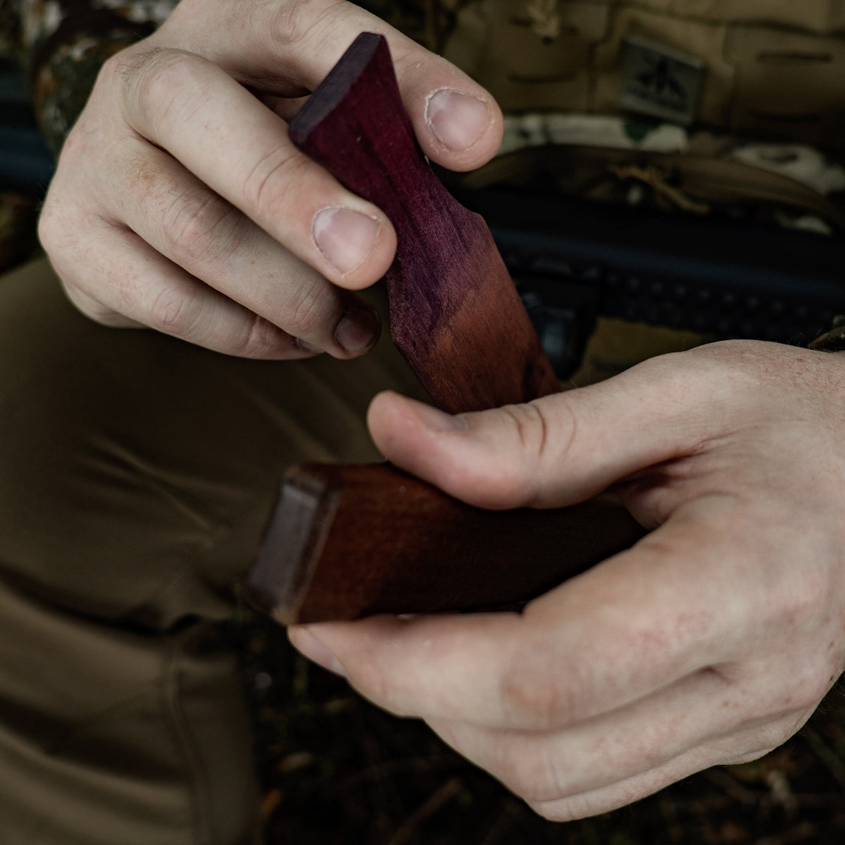 Hands hold two carved wooden firearm components: one dark purple fading to reddish-brown, the other dark brown.