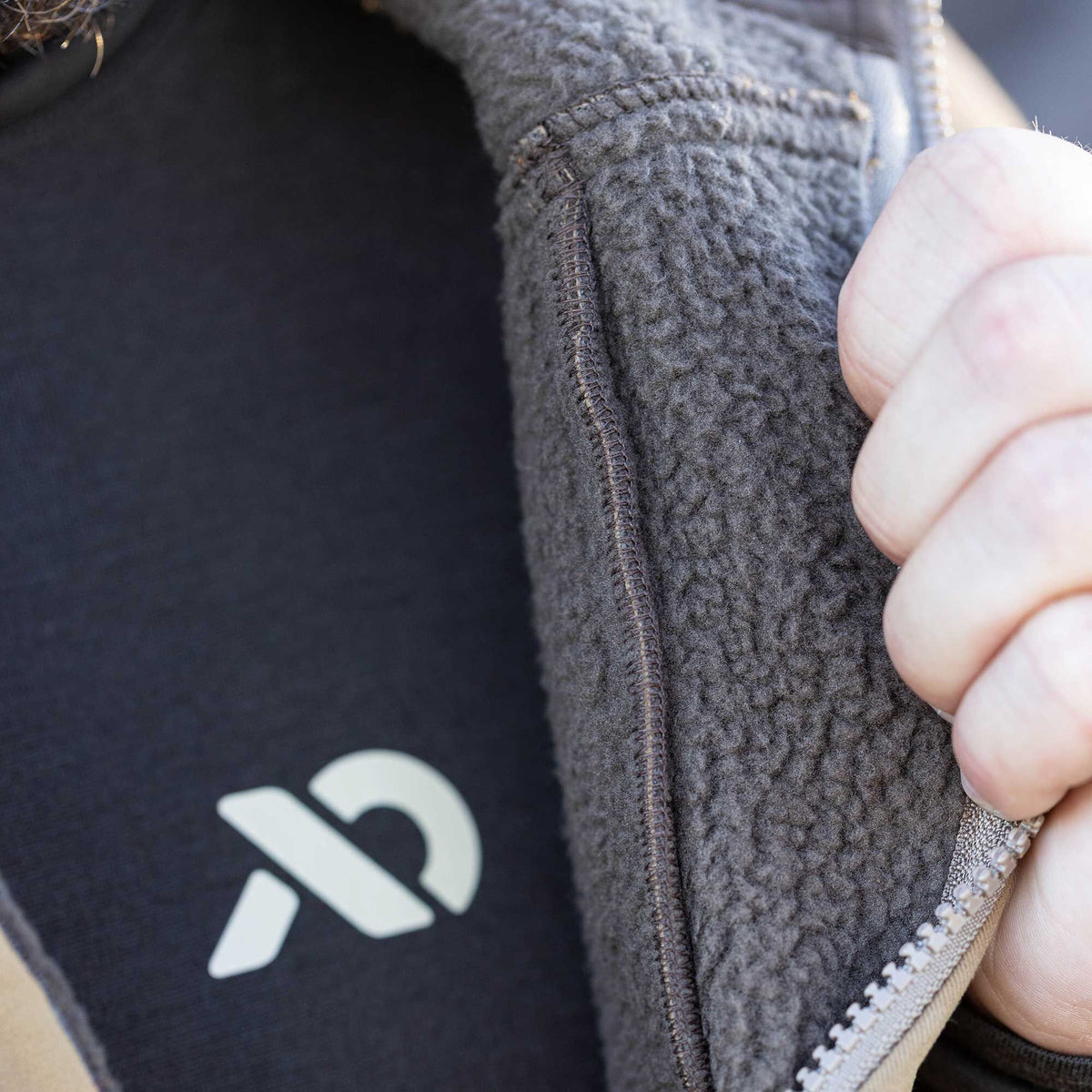 A hand pulls open a tan jacket, revealing its dark gray fleece lining and a black shirt with a light logo.