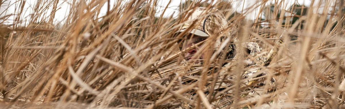 Camouflage hat & jacket on person peeking through dry reeds.