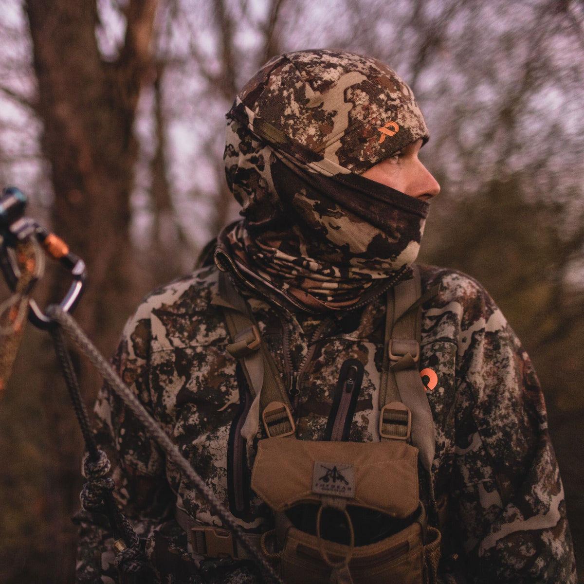 A hunter in camouflage beanie, neck gaiter, and jacket with orange logos, wearing a tan chest rig and holding a climbing rope.