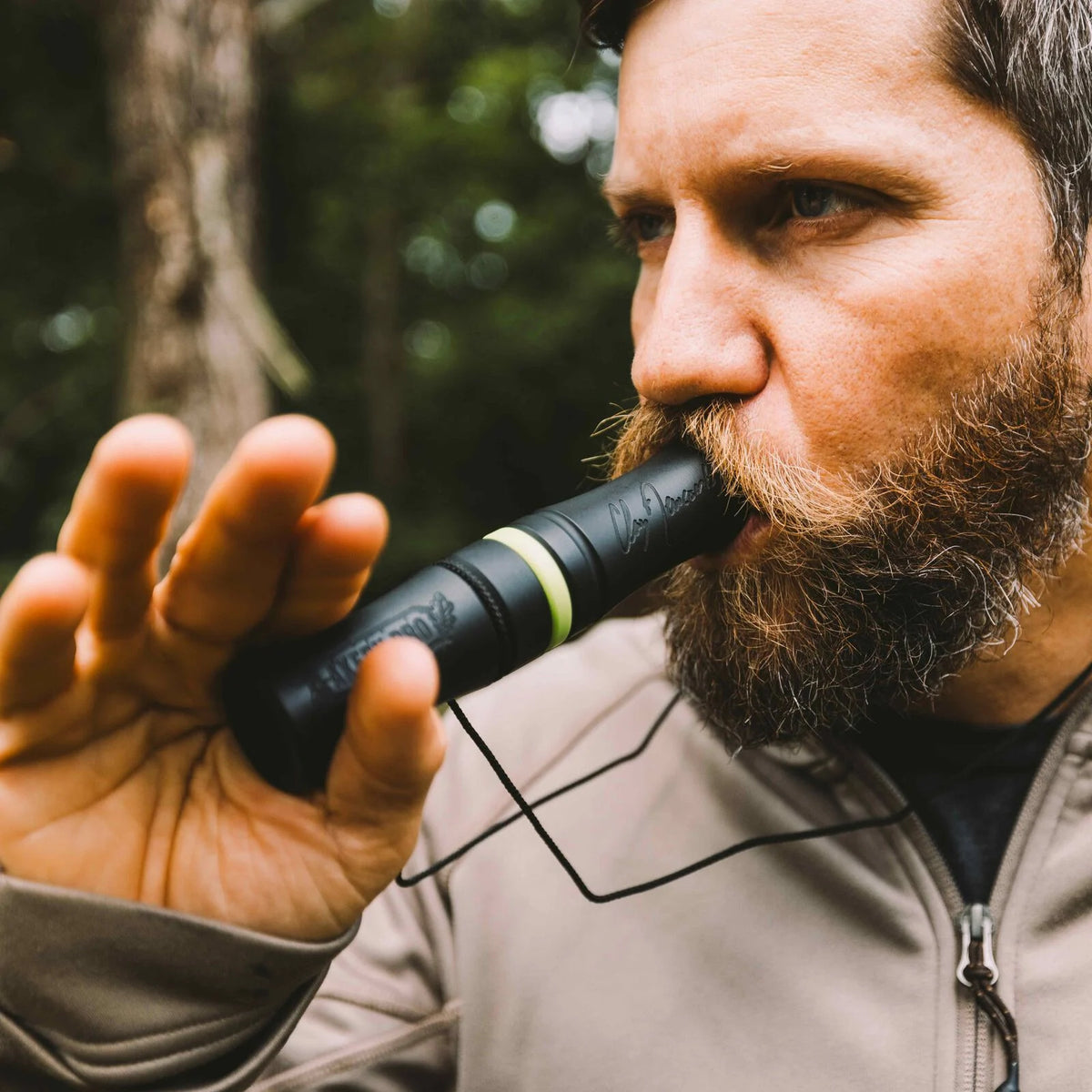 A bearded man blows a black Claw Blaster Pro game call with a bright green band, featuring a Clay Dawson signature and attached black lanyard.