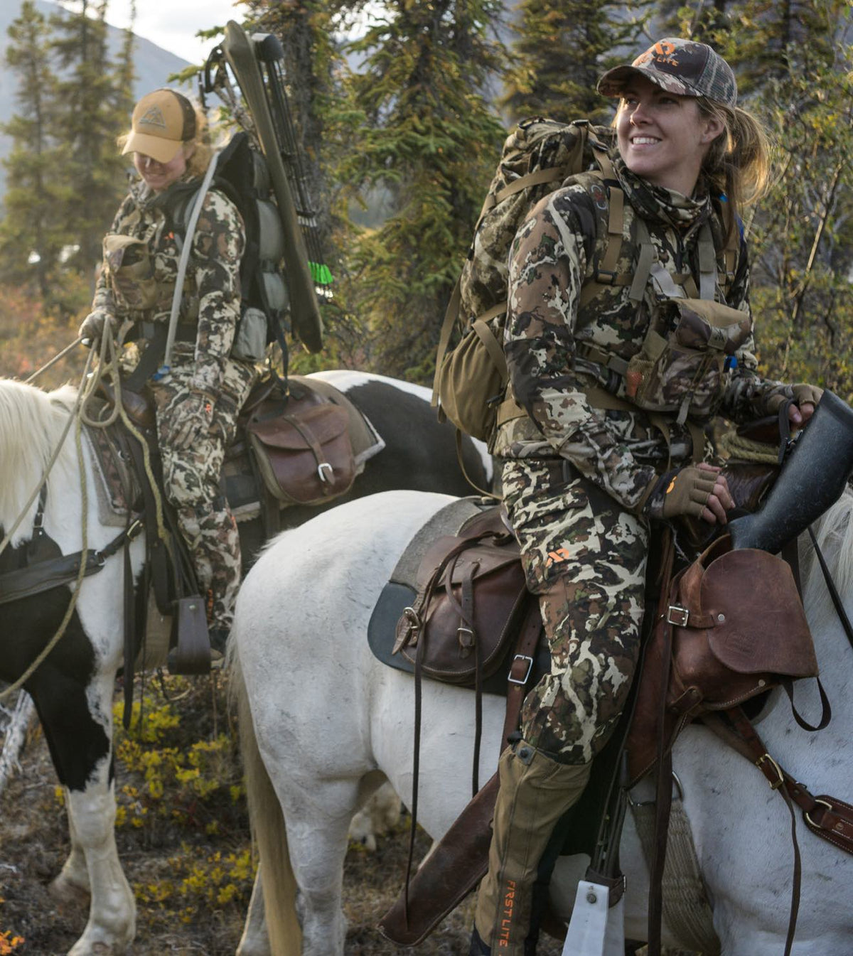Two women in camouflage hunting gear on horseback, with packs, rifle, and bow.