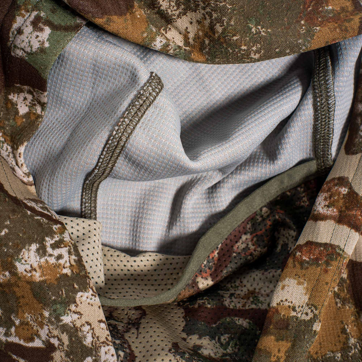 A brown, green, and tan camouflage performance top featuring a light grey grid-textured lining, perforated beige mesh, and olive green stitching.