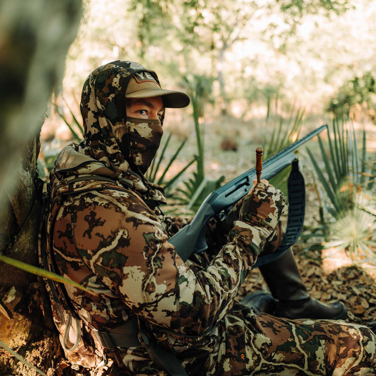Person wearing a brown, green, and tan camouflage hooded hunting suit with a mesh face mask and matching gloves, holding a shotgun.