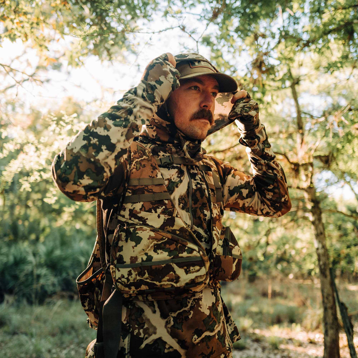 Man in multi-color camouflage hooded long-sleeve top with integrated gloves and tactical chest rig, adjusting hood.