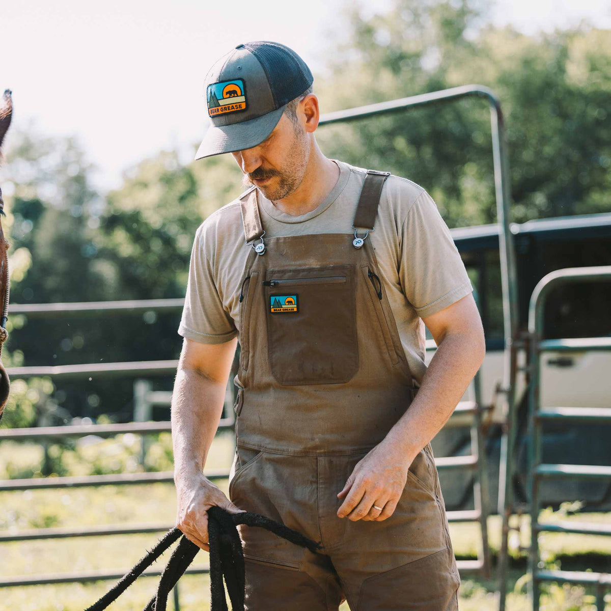 A man wearing brown work overalls over a beige t-shirt and a dark green cap with a logo patch, standing in an outdoor farm setting with metal gates and trees in the background.