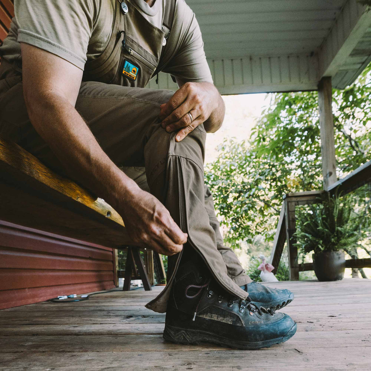A man sitting on a wooden porch adjusting his khaki cargo overalls, wearing dark hiking boots and a matching khaki shirt. The background shows a rustic wooden deck and lush green foliage.
