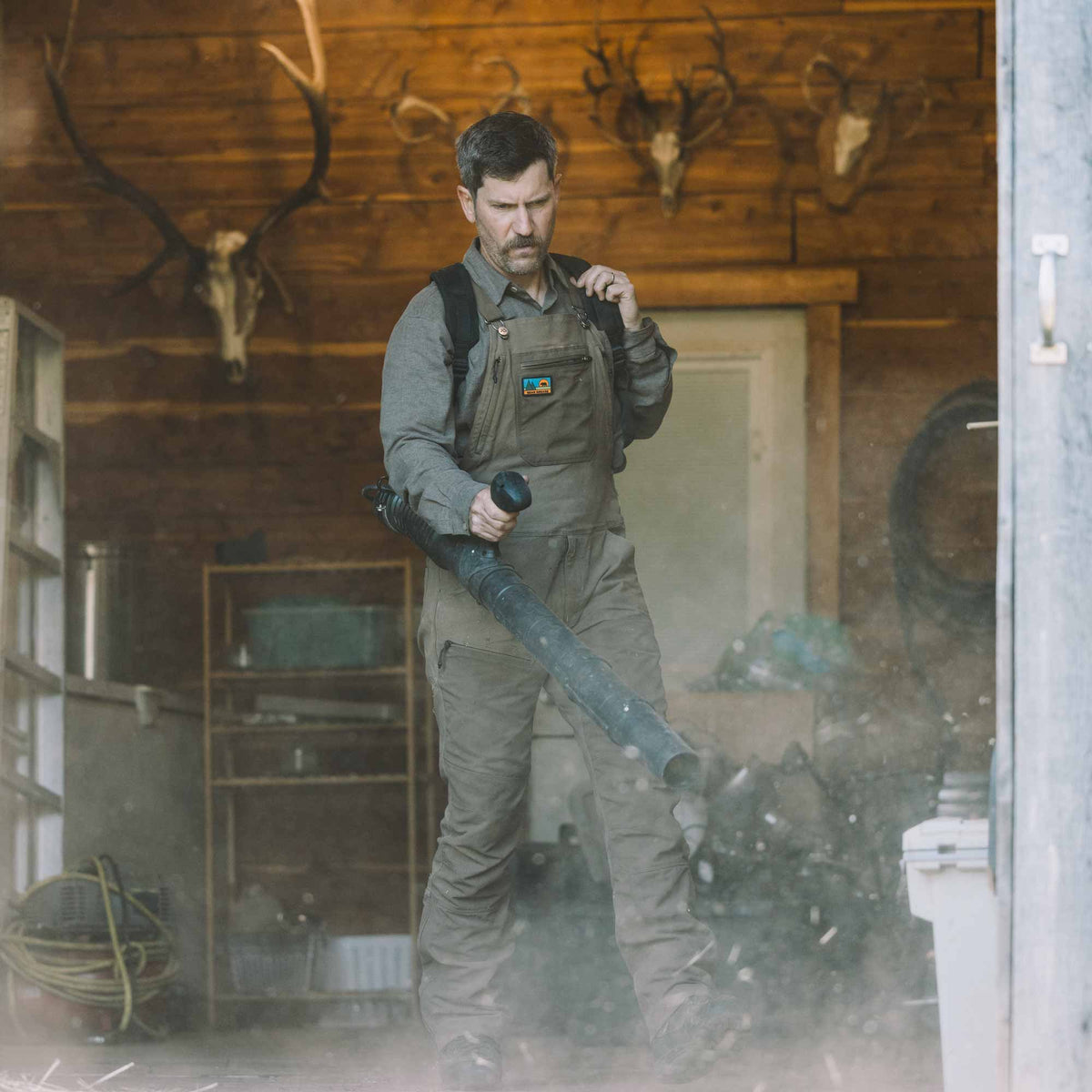 A man wearing a gray long-sleeve shirt and brown work overalls stands in a rustic wood-paneled room, directing a leaf blower toward a pile of leaves. Antlered and skull mounts are visible on the wooden walls in the background.