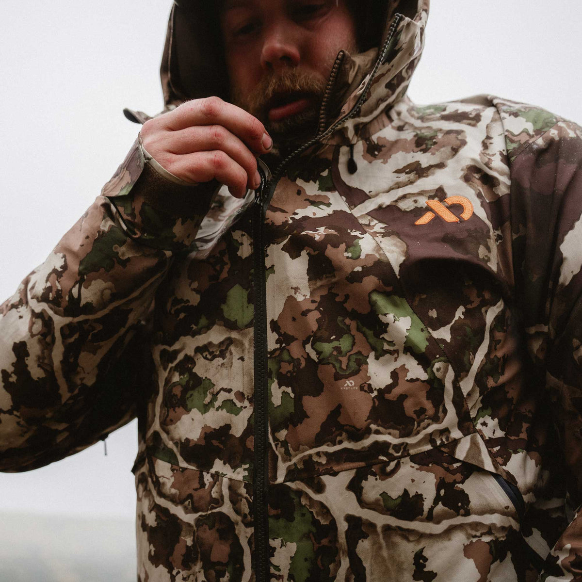 A man wearing a hooded camouflage rain jacket with a brown and green camo pattern and an orange logo on the chest, adjusting the zipper.