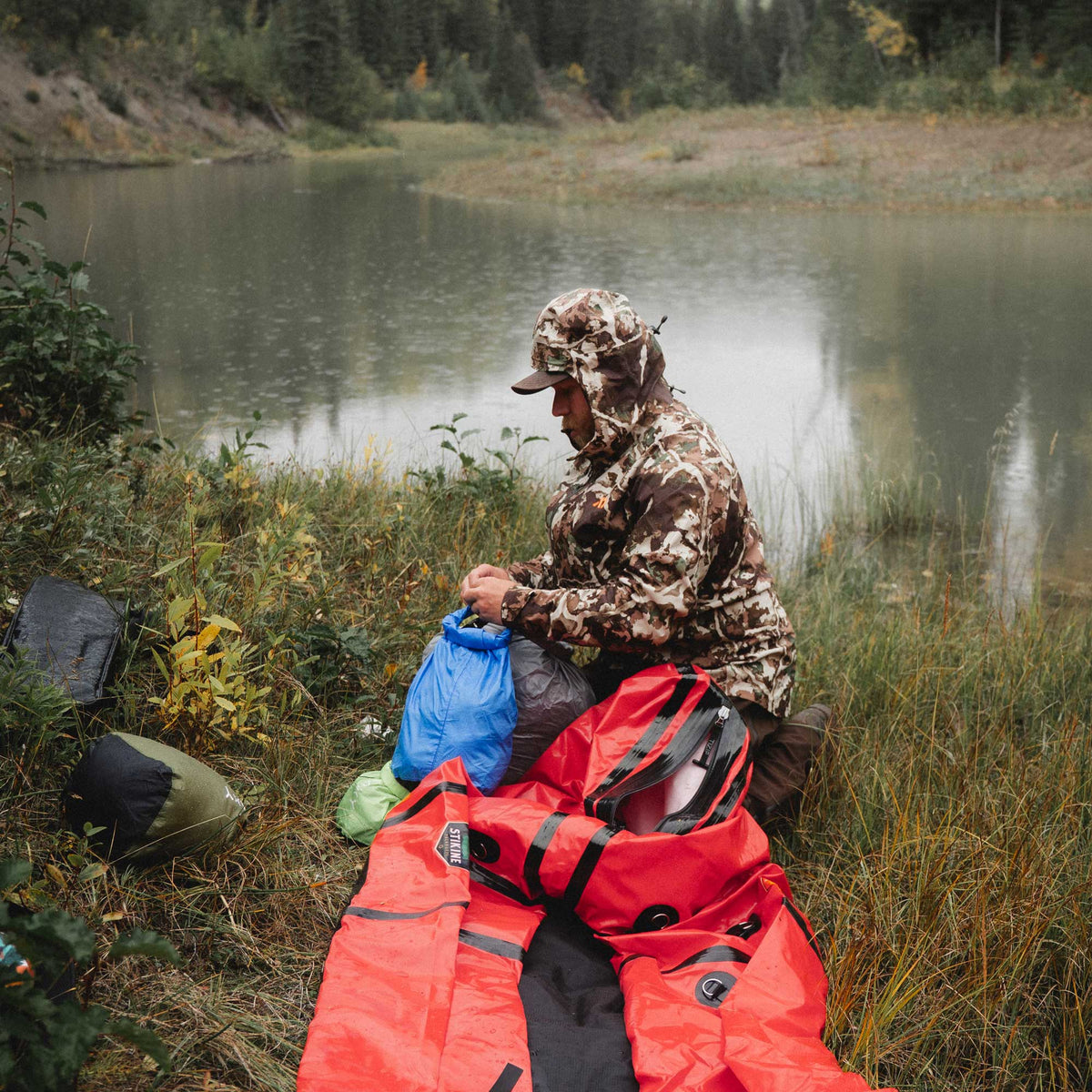 A person in a camouflage hooded jacket sits by a lake outdoors, handling a bright red inflatable kayak with black straps and a blue bag nearby. Other gear items are on the grass, with trees and water in the background.