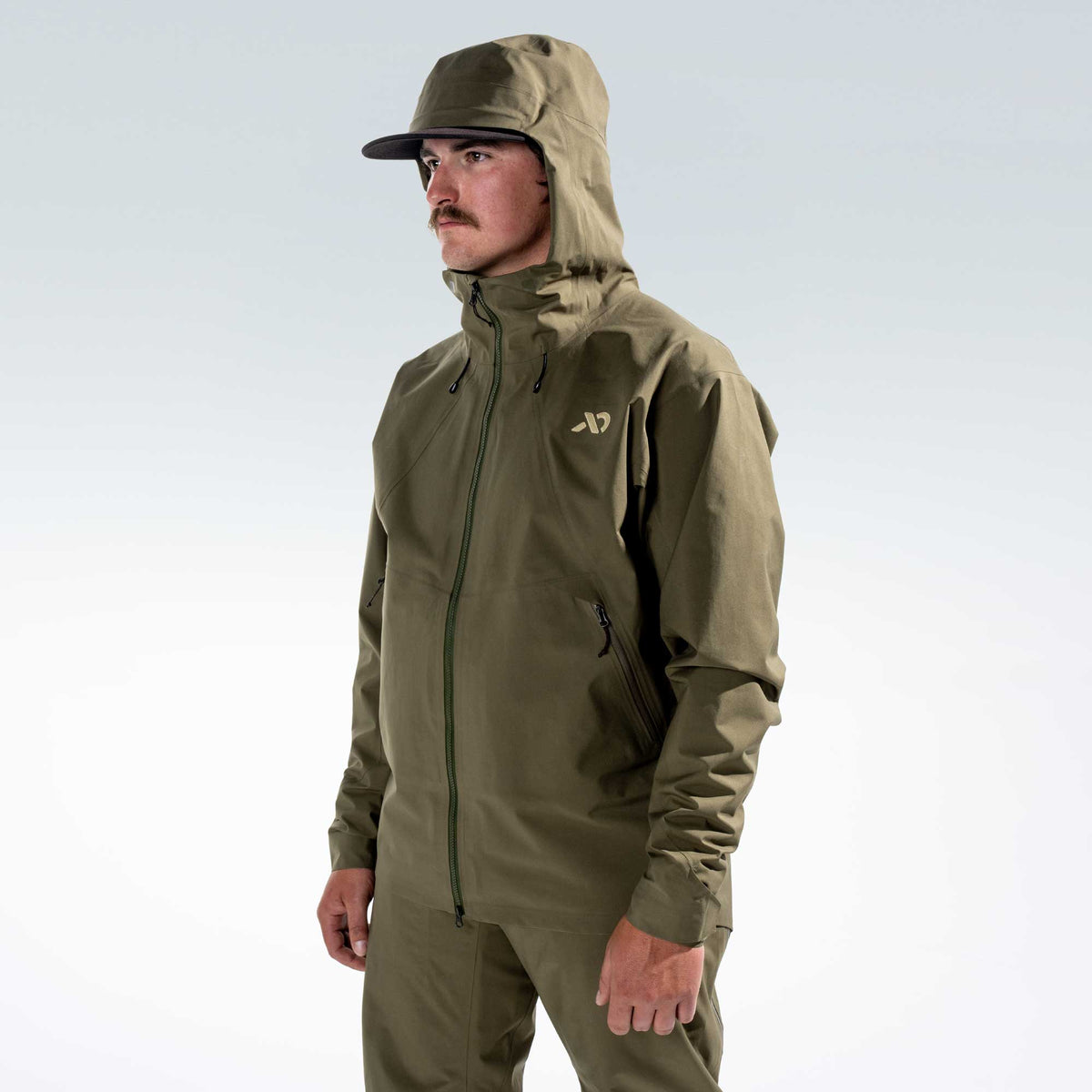 A man wearing an olive green hooded rain jacket and matching pants, standing against a light gray studio background. The jacket has a small white logo on the left chest and a zippered front.
