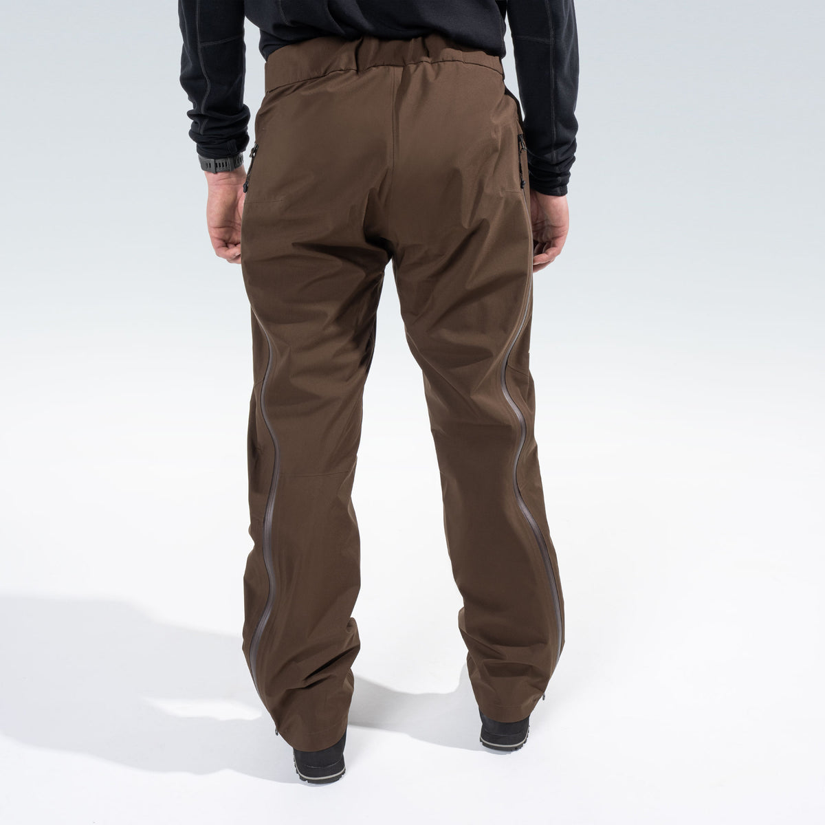 Rear view of a person wearing brown technical pants with side zips, paired with a black long-sleeve top, against a light gray studio background.