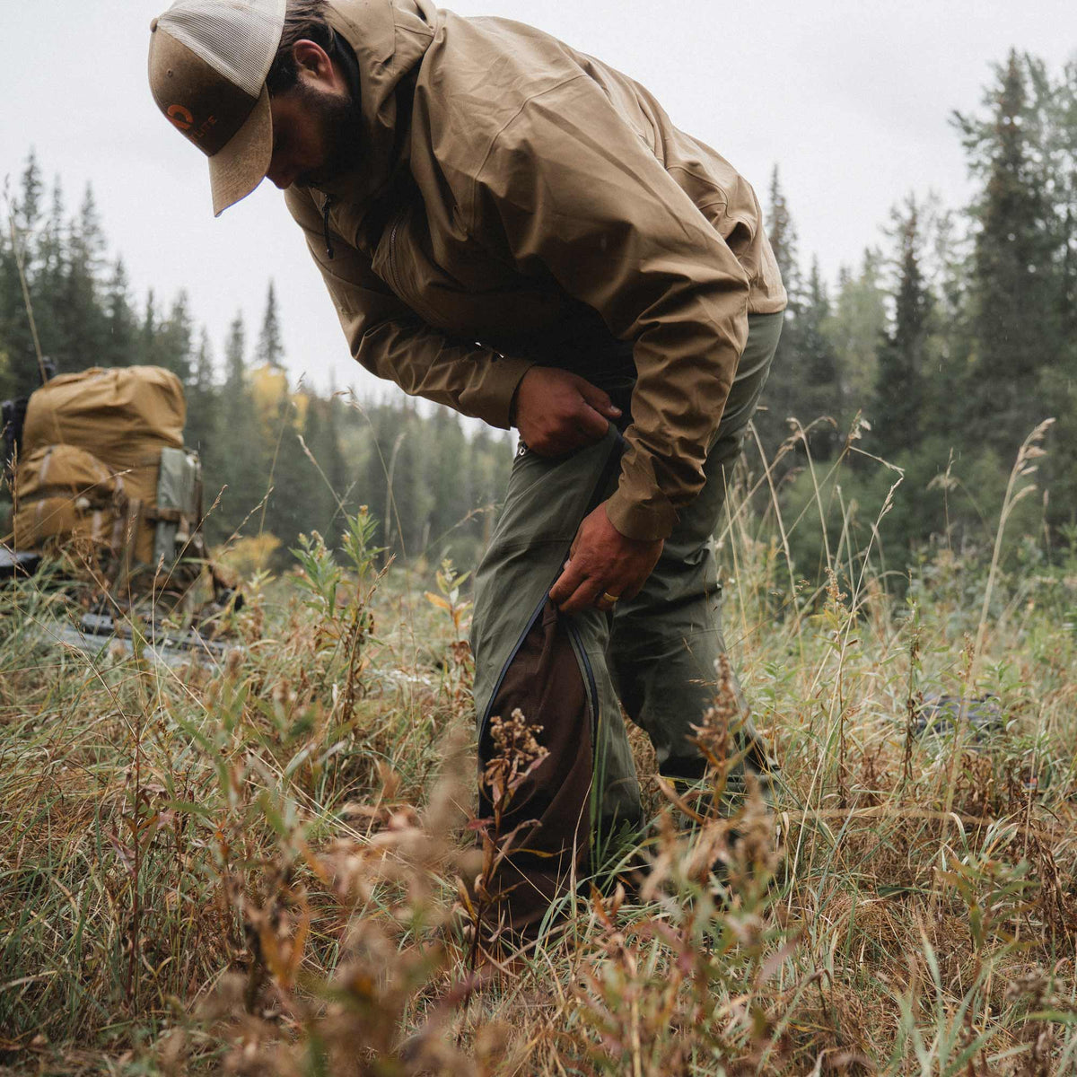 A man in outdoor gear bends down in a grassy forest clearing, zipping up green rain pants. He is wearing a tan jacket, green pants, and a beige cap, with a hiking backpack visible in the background among pine trees.