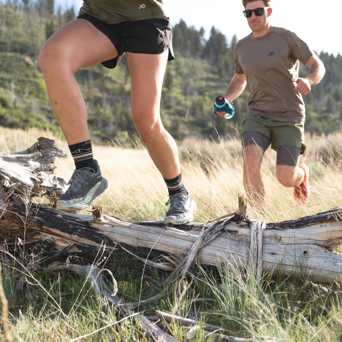 Two people running through a forest with one person stepping over a log.