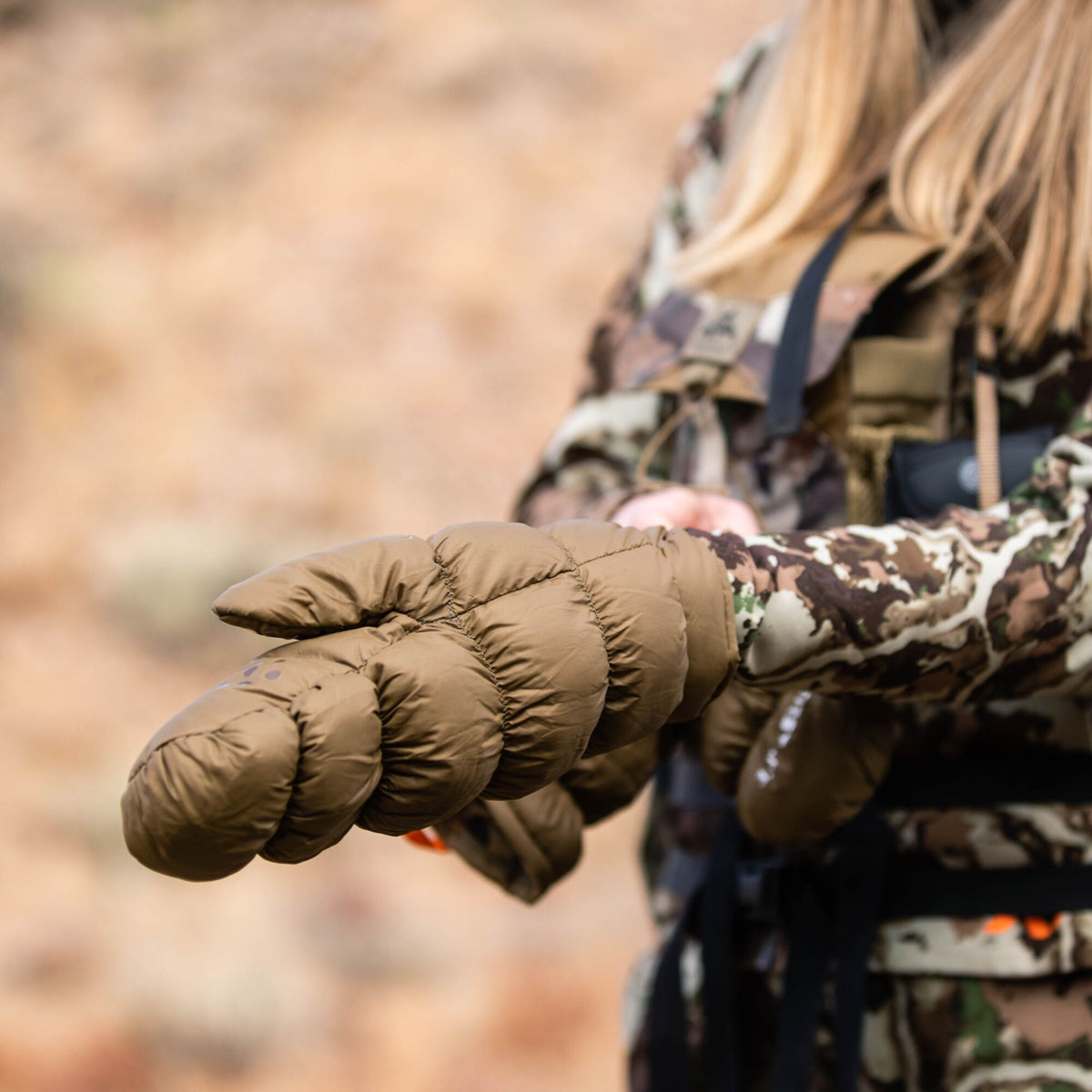 Olive brown puffy insulated mitten with a dark dot pattern on the thumb worn by a person in camouflage.