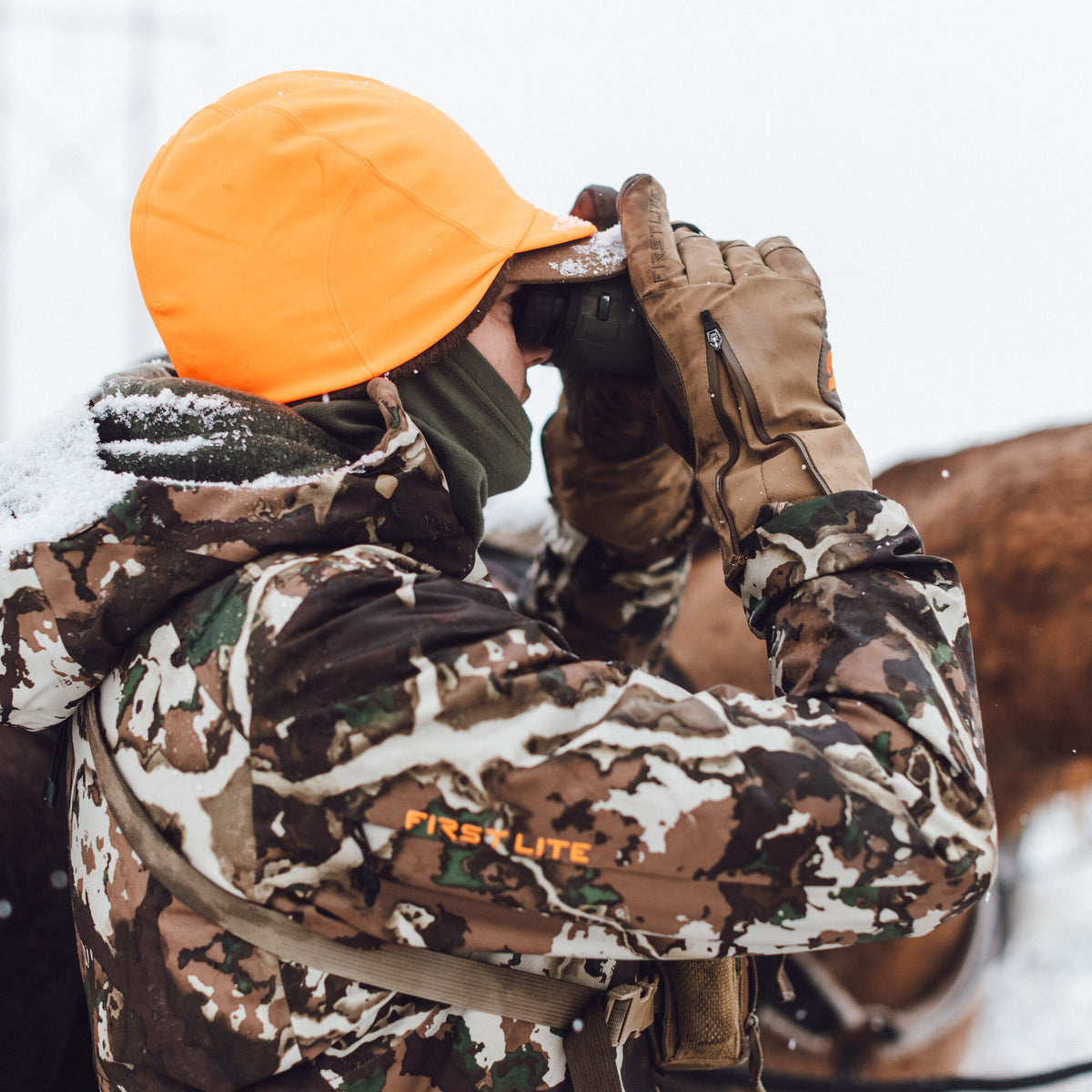 First Lite hunter in an orange beanie, camouflage jacket with orange logo, and tan gloves looks through binoculars in the snow.