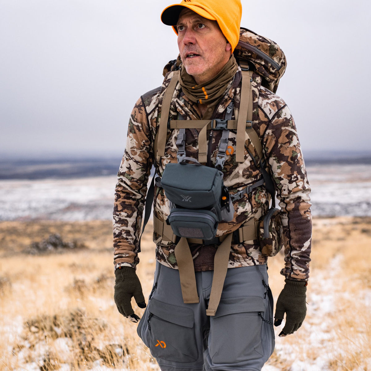Hunter in camouflage long-sleeve shirt, dark grey cargo pants, and orange beanie wears a dark grey Vortex binocular chest harness and camouflage backpack in a snowy field.