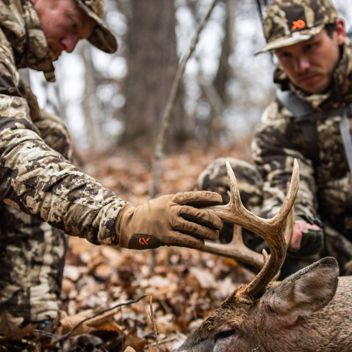 Tan FIRST LITE hunting gloves with an orange logo grip deer antlers, worn by a hunter in camouflage.