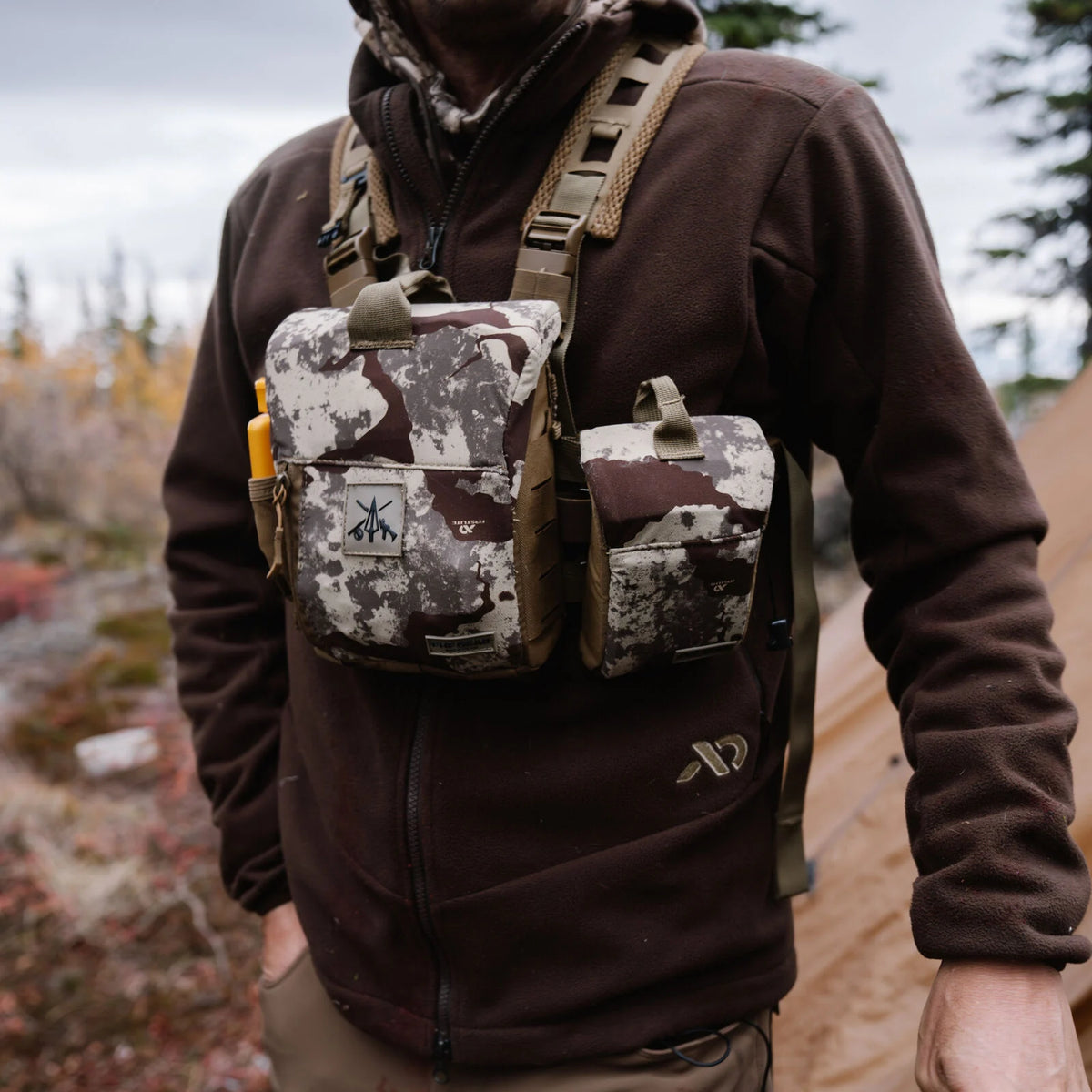 A brown, white, and grey camouflage chest rig with a main pouch displaying a logo, a smaller side pouch, and tan shoulder straps, featuring a yellow item in its side slot.