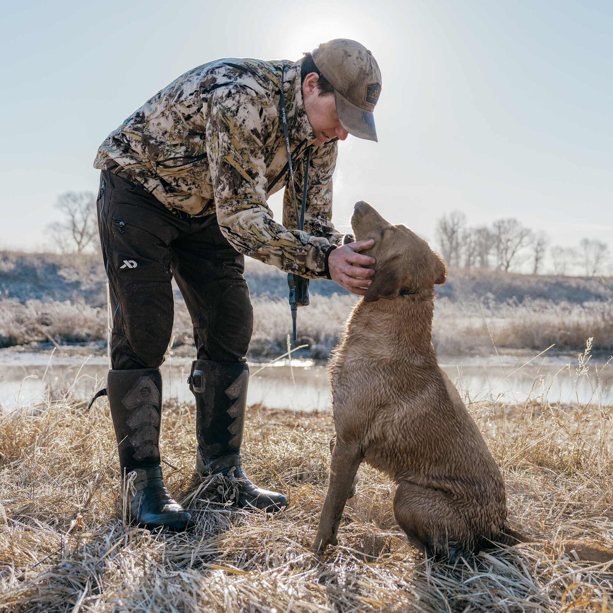 Man wearing camouflage jacket, dark brown technical hunting pants with side zippers and a white logo, and tall black rubber boots, petting a wet dog by a frosted pond.