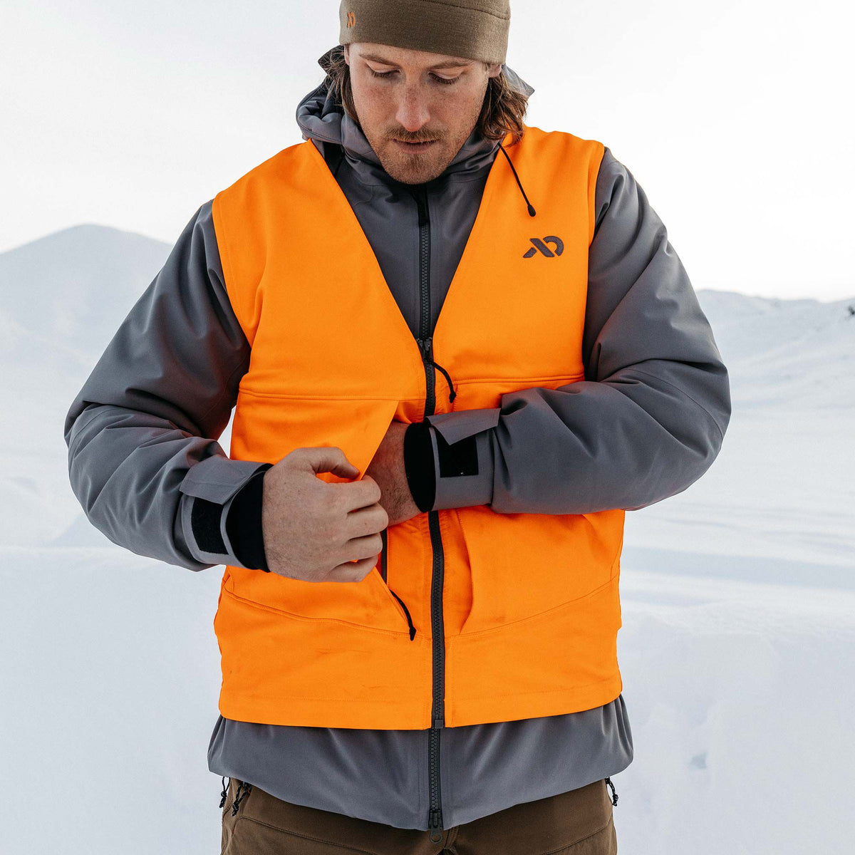 Hunter orange zippered vest with a black chest logo worn over a grey jacket, being adjusted by a person in the snow.