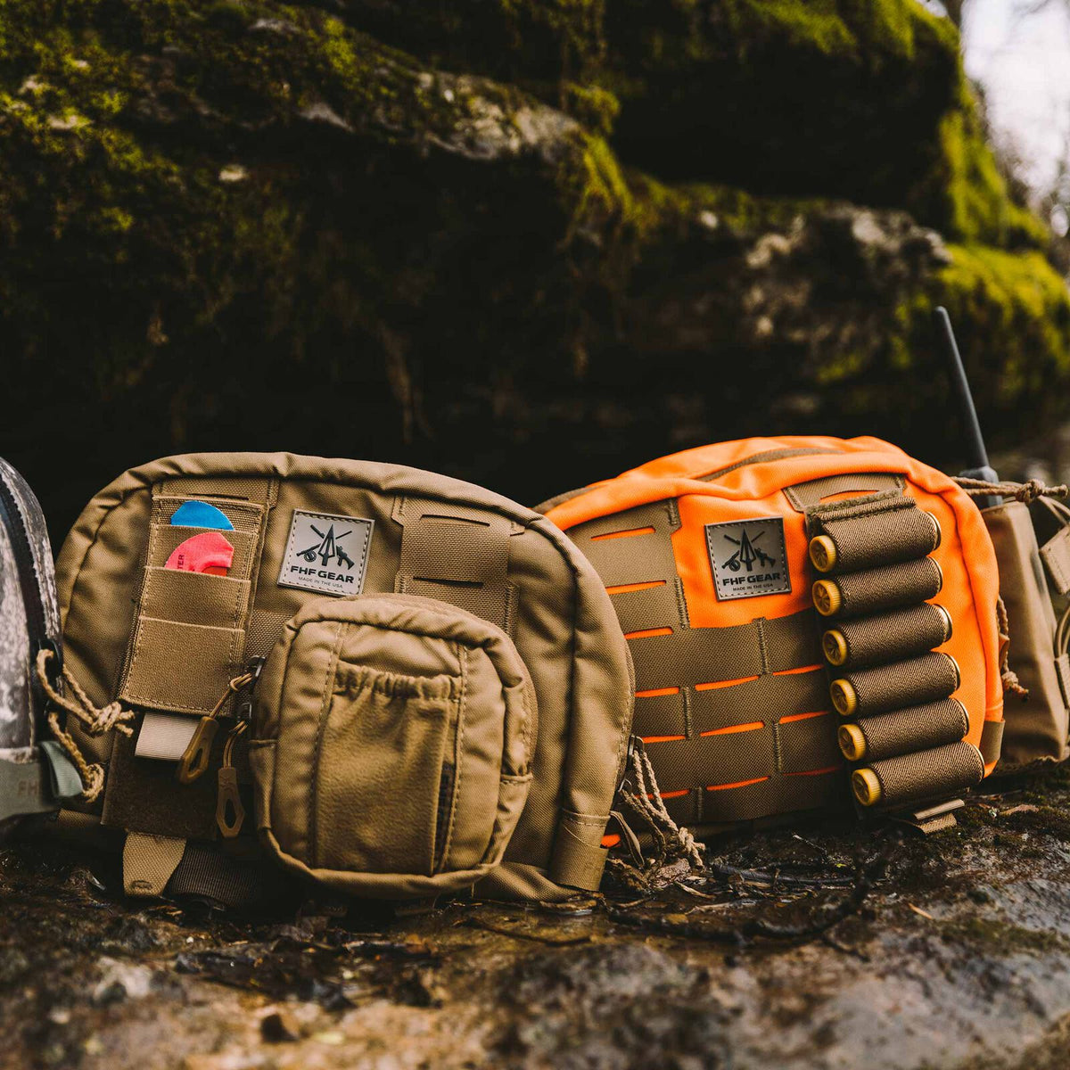 Two FHF GEAR modular gear pouches, one coyote brown with an attached small pouch and the other high-visibility orange with shotgun shell holders and MOLLE webbing, rest on mossy ground.
