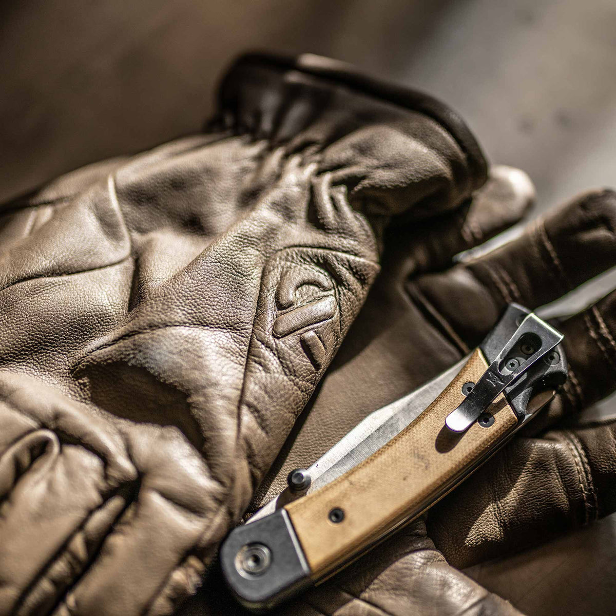 Tan-handled folding knife with a silver blade and pocket clip resting on brown leather gloves with an embossed logo.