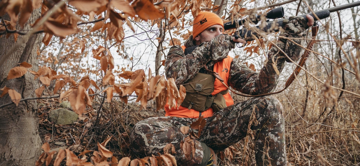Hunter wearing orange beanie, vest, camo, and olive chest rig aiming rifle.