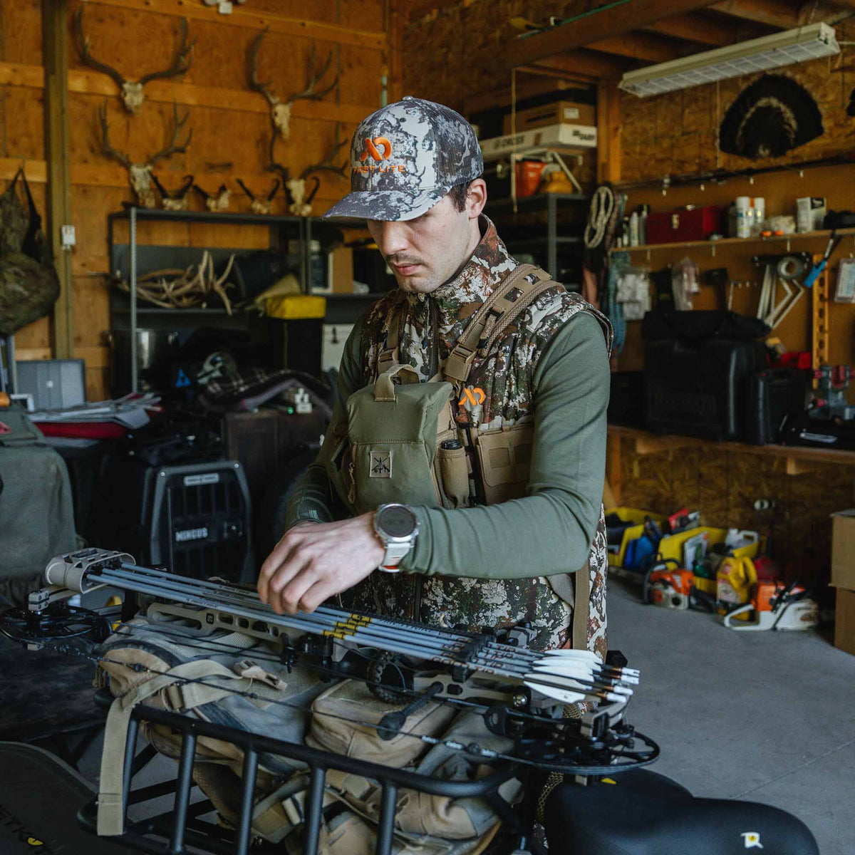An olive green hunting chest rig with tan straps, worn over a camouflage vest by a man adjusting a bow and arrows.