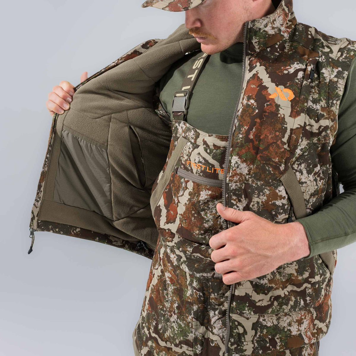 Man in a multi-color camouflage vest with an orange logo and bibs, revealing the vest's olive green fleece interior with an internal mesh pocket, and "FIRST LITE" text on the bib pocket.