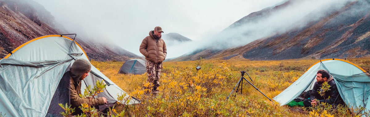 Three grey camping tents among men, yellow shrubs, and foggy mountains.