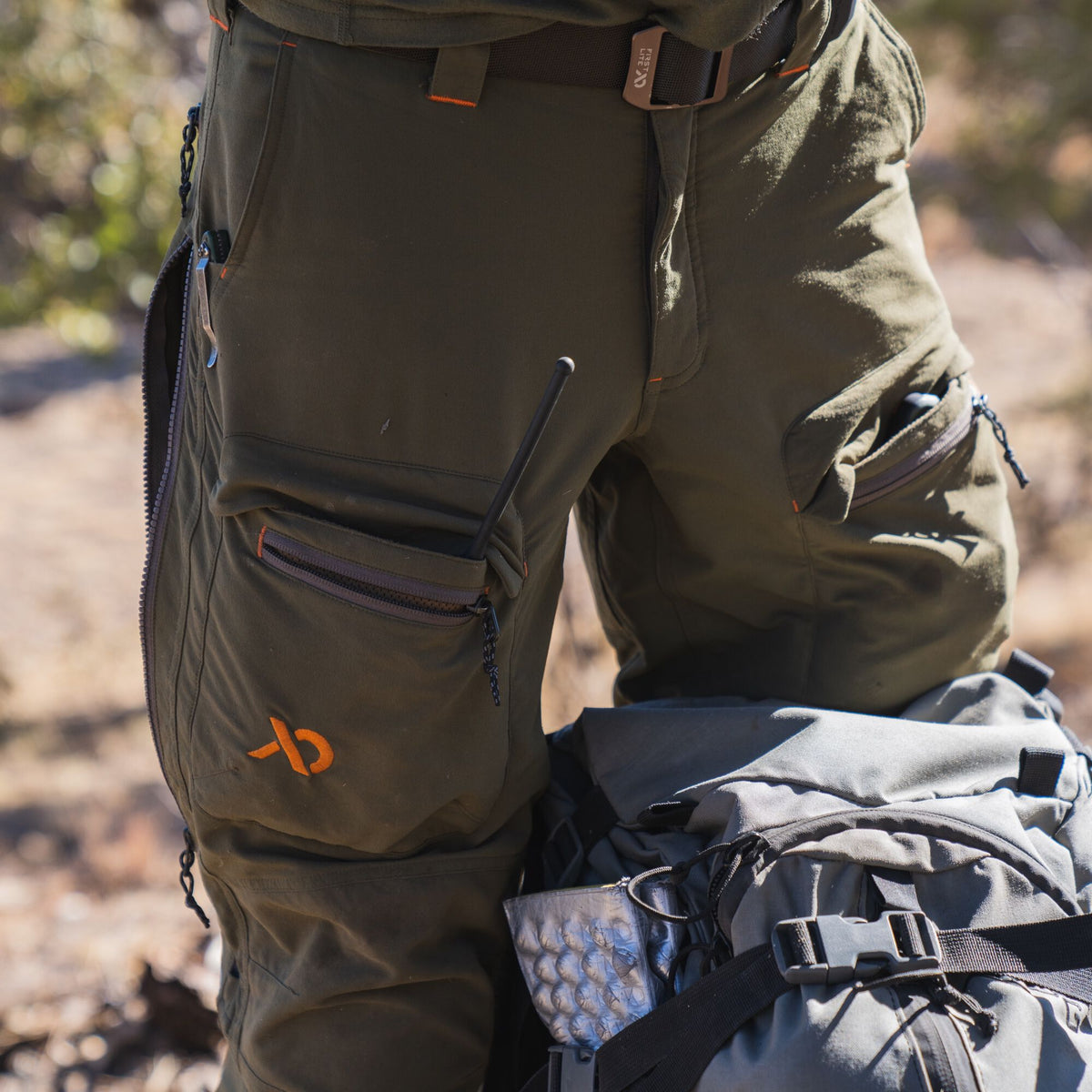Dark olive green cargo pants with zippered pockets, an orange logo, orange stitching accents, and a side leg zipper.