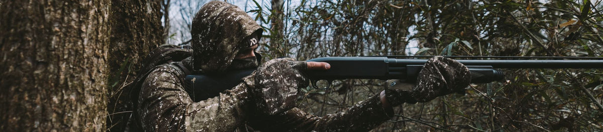 Camouflage-clad person in a hooded jacket and gloves aims a black shotgun.