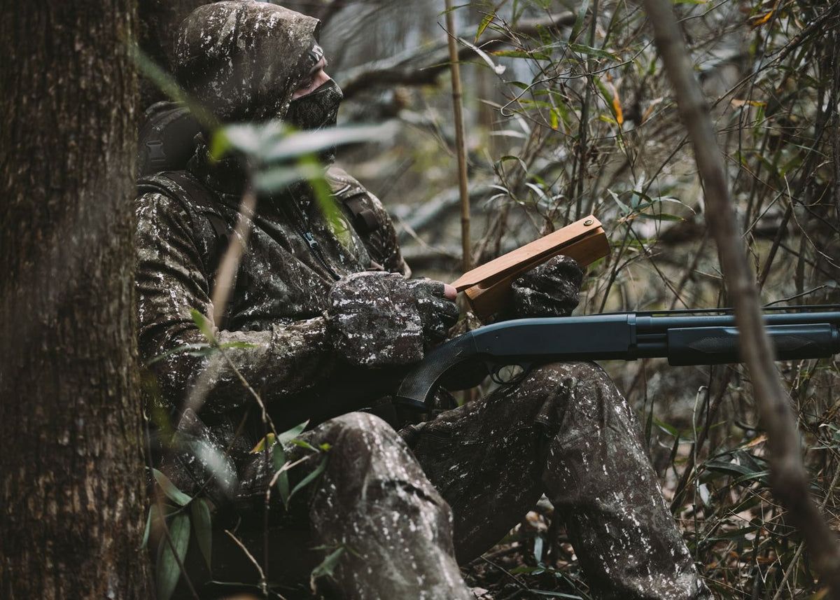 Camouflage-clad hunter with shotgun and wooden box call, hooded and masked in forest.
