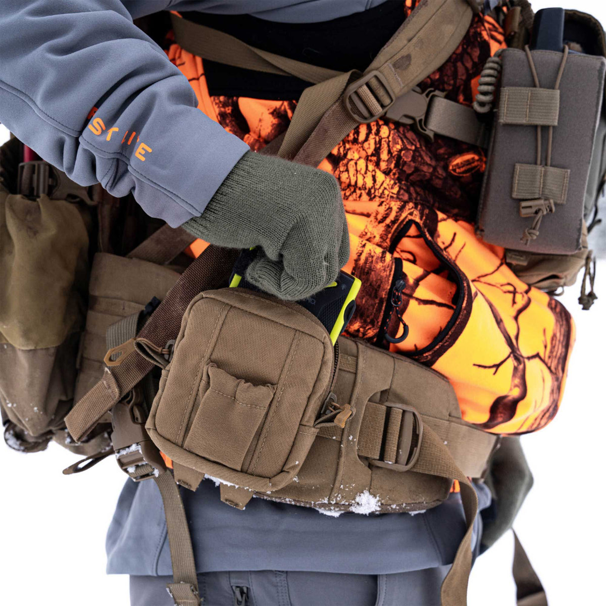 A person wearing a gray jacket with 'FIRST LITE' on the sleeve and olive gloves over an orange camo top and tan modular vest, accesses a black and yellow device from a tan pouch.