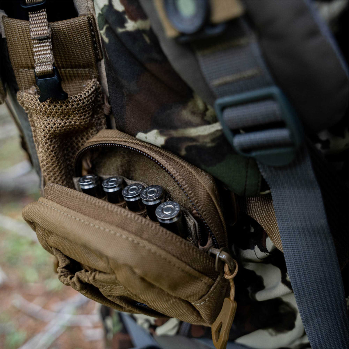 Coyote brown tactical pouch attached to a camouflage backpack, showing four cartridges in its open zippered compartment and a mesh side pocket.