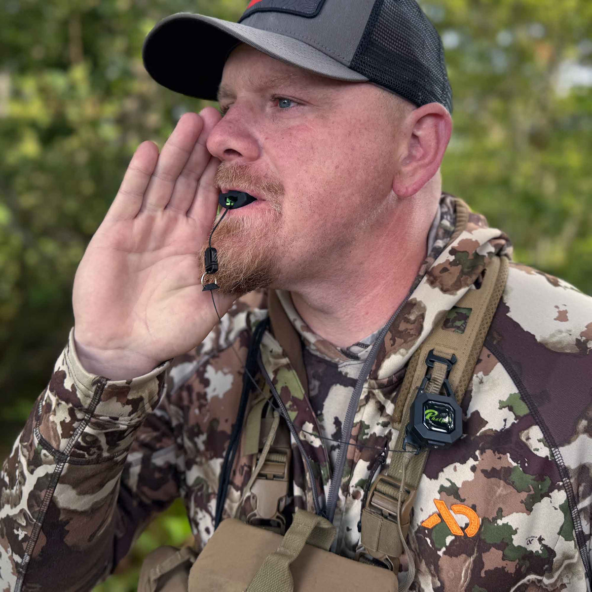 Man in camouflage using a black and green hunting call device held in his mouth, with a second black device clipped to his chest strap.