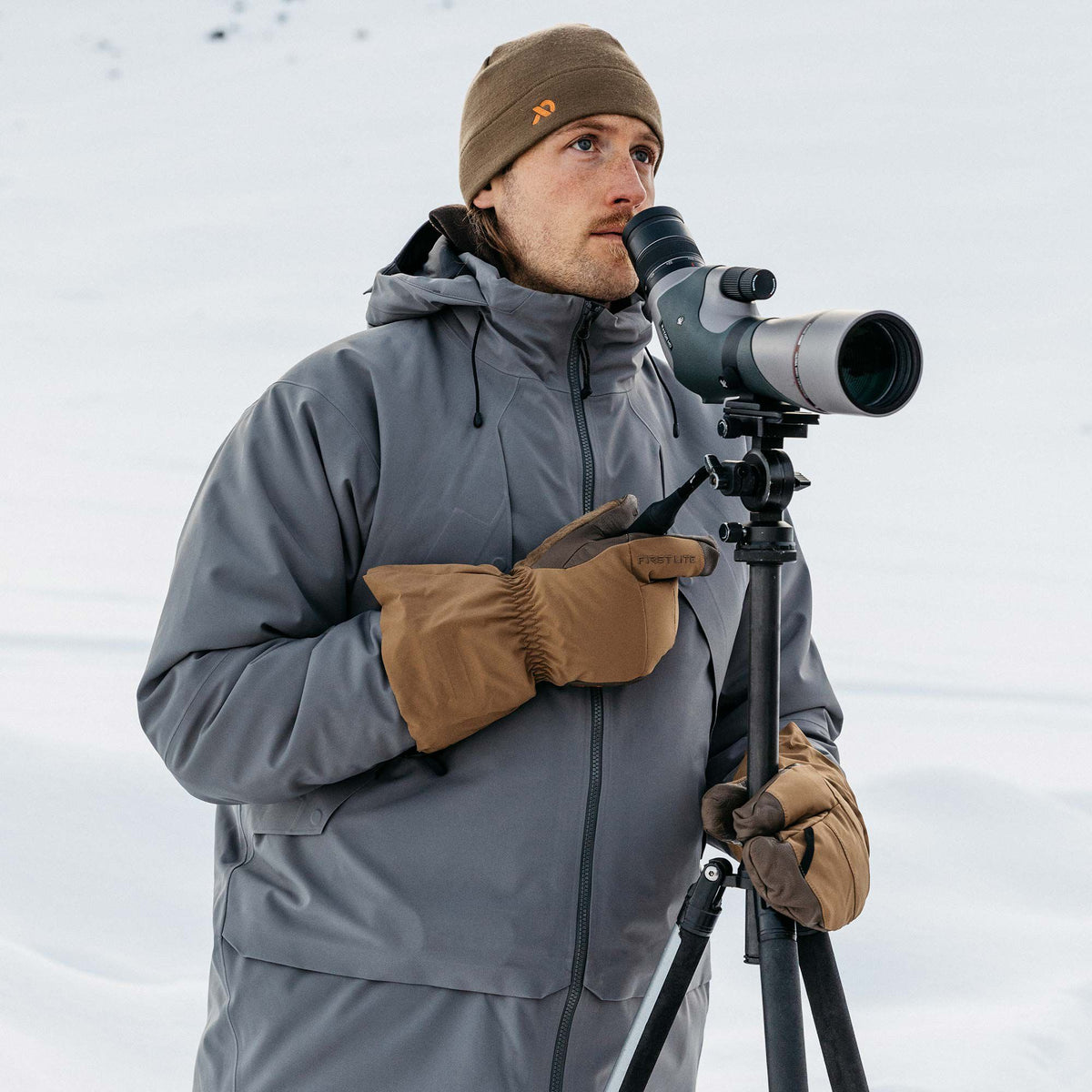 A man wearing a grey hooded jacket, brown insulated gloves with a FIRSTLITE logo, and a brown beanie looks through a spotting scope in a snowy environment.