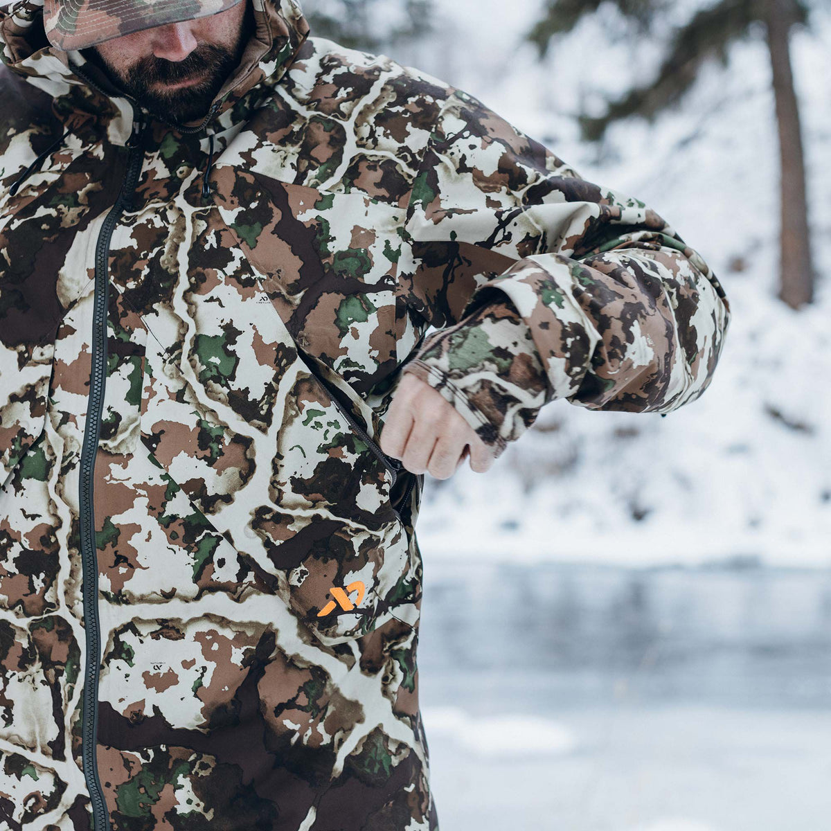 Hooded brown, green, and white camouflage jacket with an orange logo and zippered pocket, worn by a person zipping it.