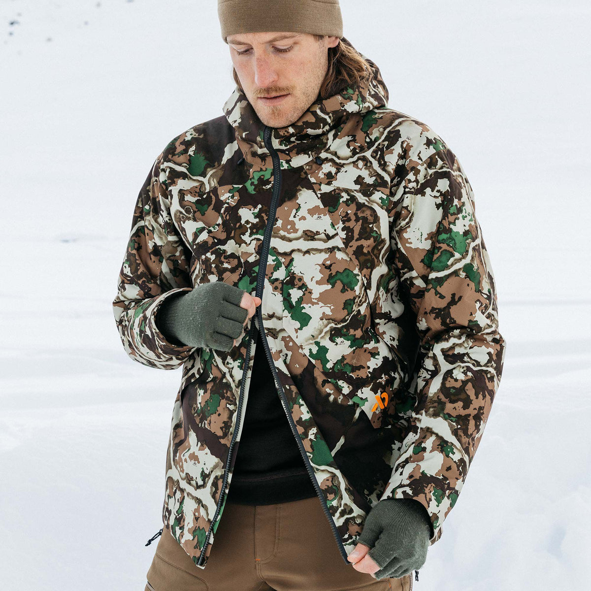 A man wears a hooded camouflage jacket in brown, green, and white, with an orange logo, unzipping it in the snow.