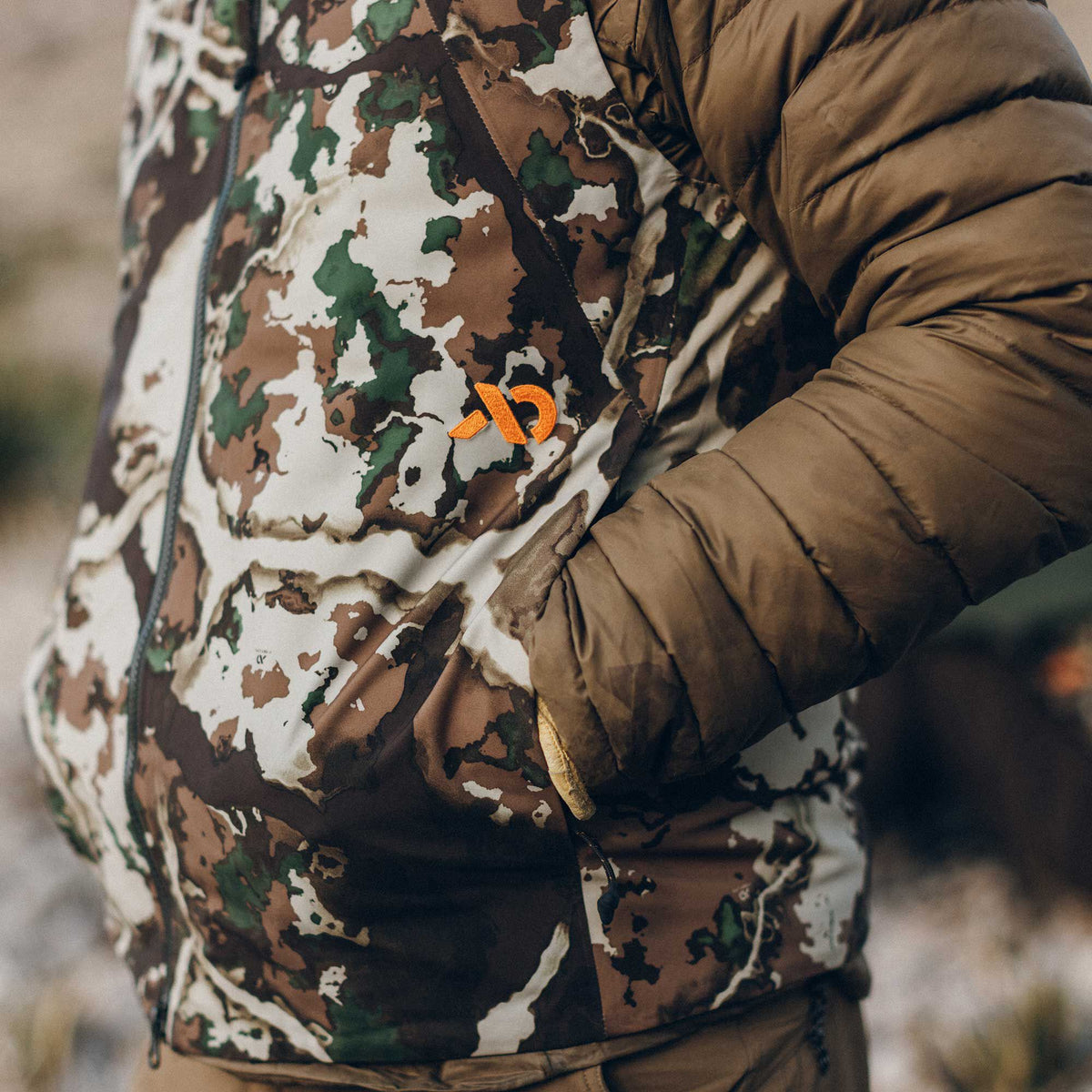 Camouflage jacket with brown, green, and cream pattern, solid brown puffy sleeves, and an orange embroidered chest logo.
