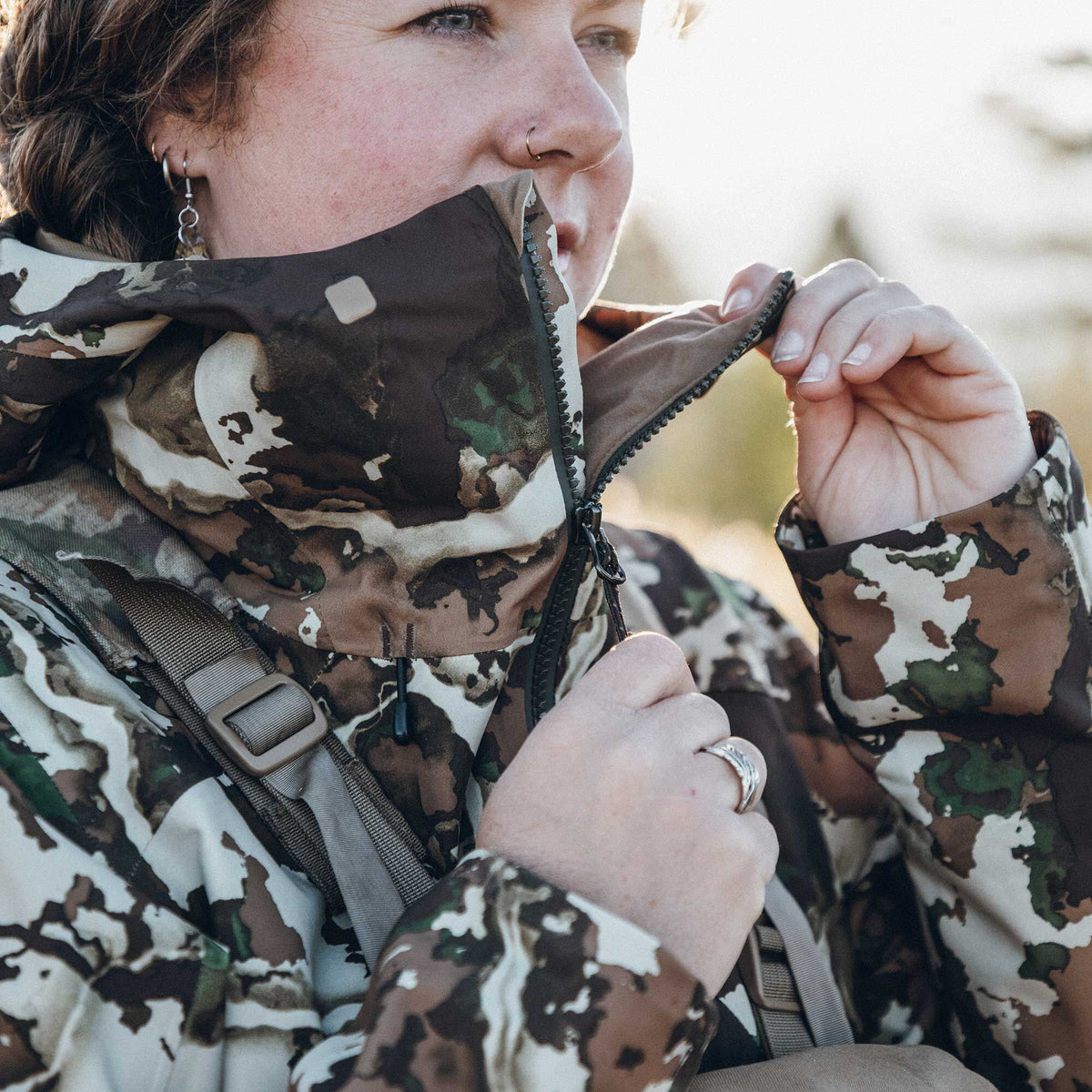 Person zipping up a brown, white, and green camouflage jacket with a high collar and visible backpack strap.