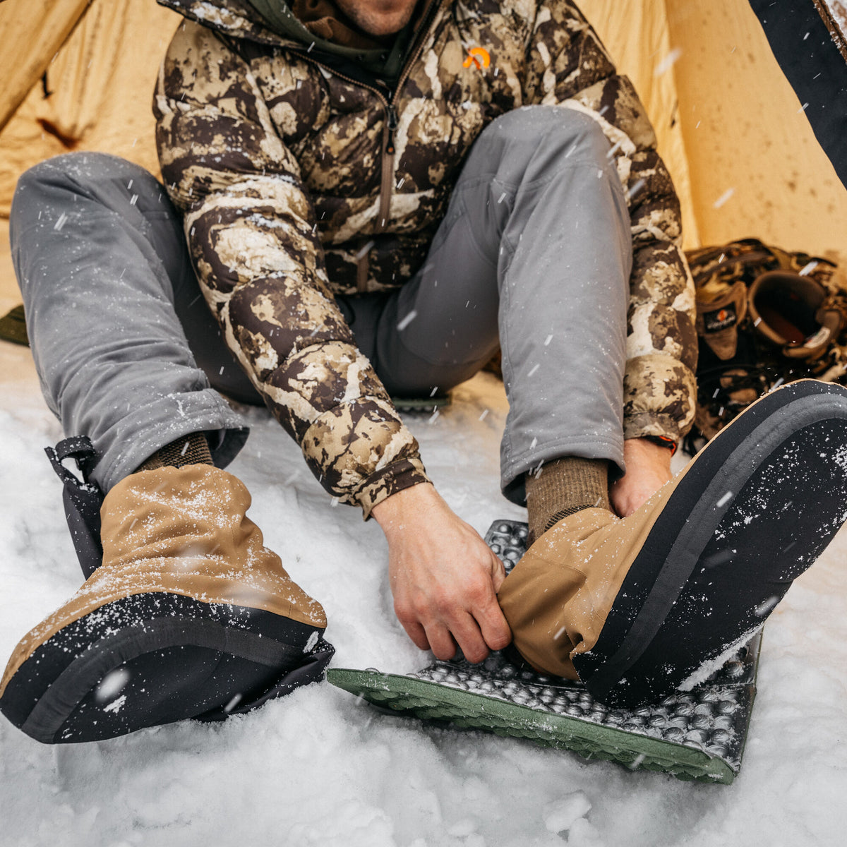 Tan insulated camp booties with black soles are worn by a person adjusting one in the snow.