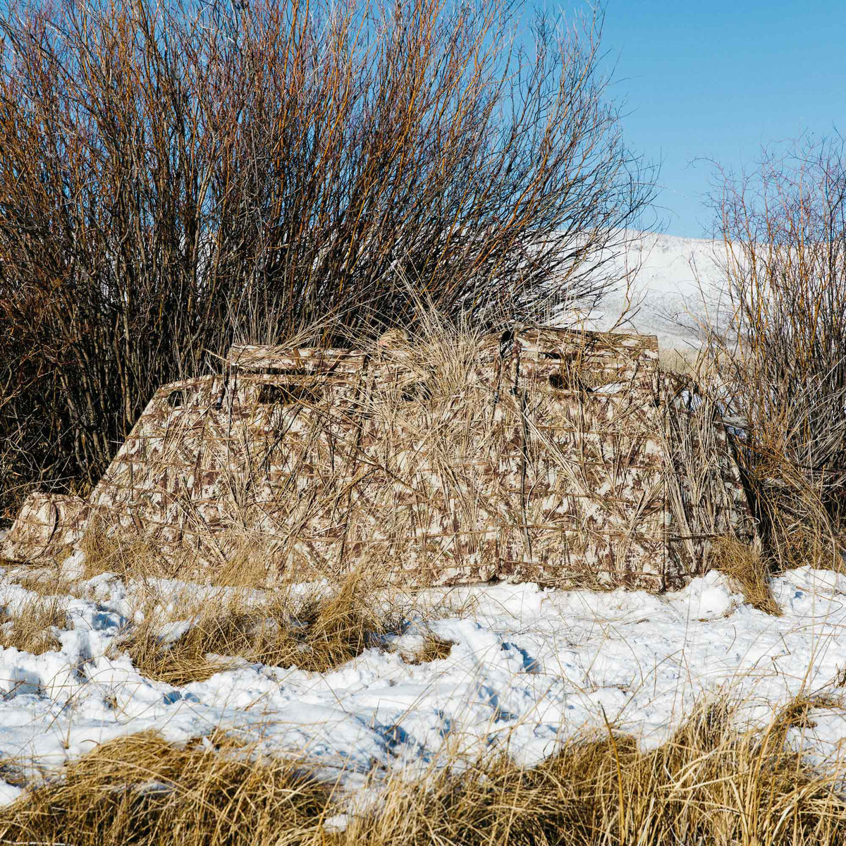 Camouflage hunting blind in a snowy, reedy field, featuring brown, tan, and white patterns augmented with natural reeds.