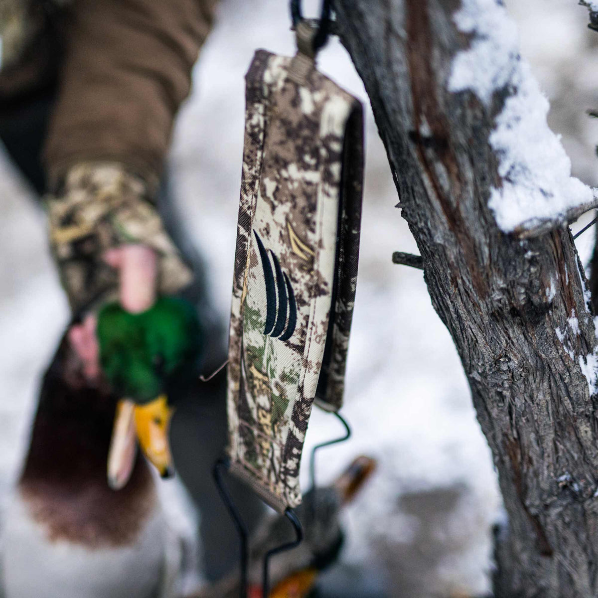 Camouflage hunting lanyard with a black claw logo, hanging on a snowy tree branch, with a hunter holding a duck blurred in the background.