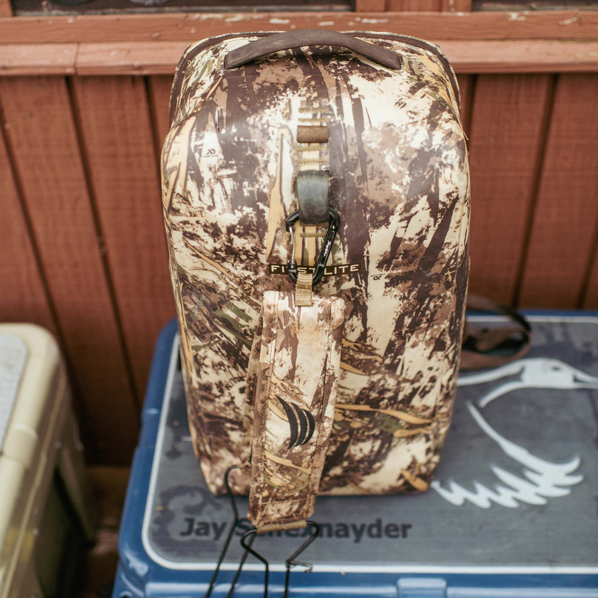 First Lite brand camouflage ground control with a top handle, an attached carabiner, and a side strap displaying an emblem.